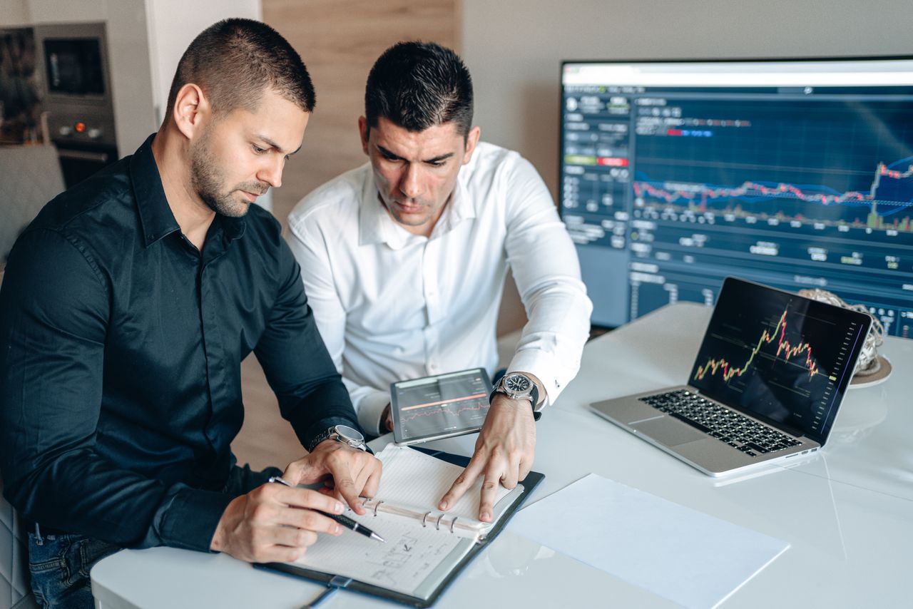 services-04 Two businessmen analyzing financial charts on digital devices in a modern office setting.